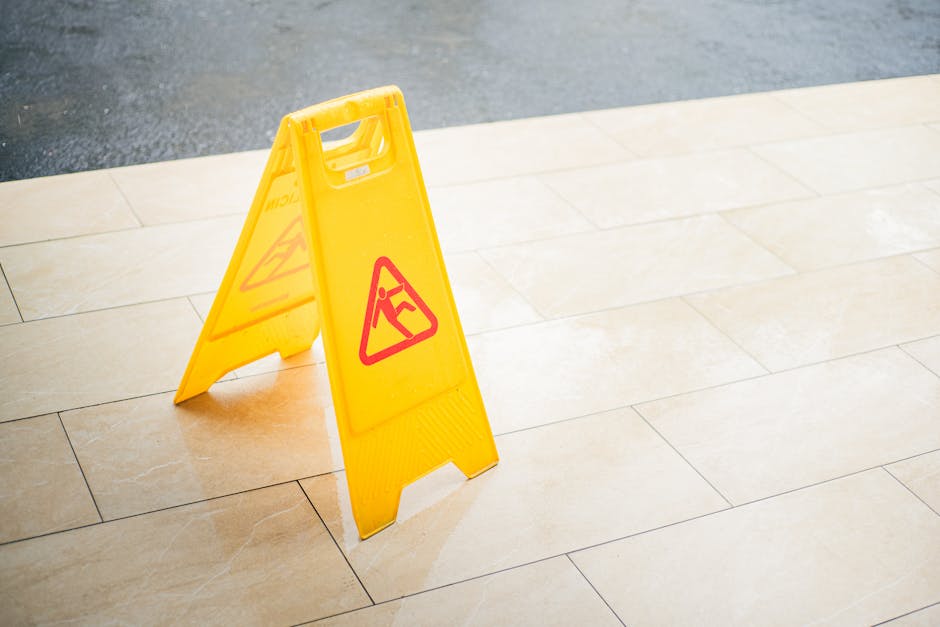 A yellow plastic warning sign with red hazard symbols stands on a polished beige tiled floor, indicating a caution or wet surface area. The sign is positioned near a clean, well-lit space with light r
