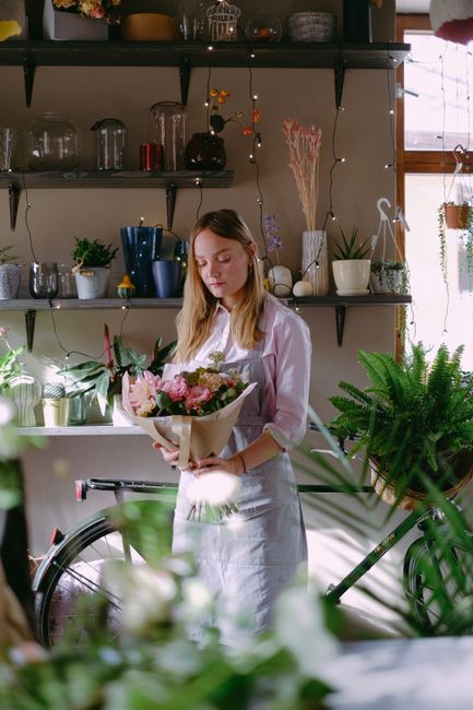 A young woman with long blonde hair, wearing a white shirt and apron, is holding a bouquet of fresh flowers wrapped in light brown paper. The bouquet features pink roses, light purple flowers, and lus