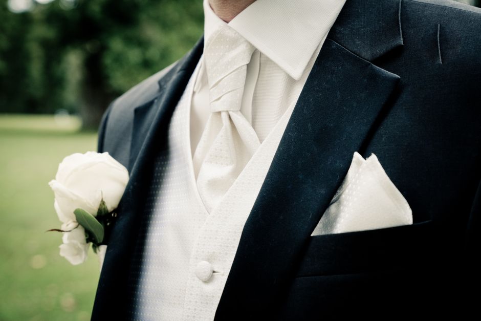 Close-up of a groom wearing a classic black tuxedo with a white dress shirt and a pale beige cravat, holding a boutonnière consisting of a single white rose with green leaves pinned to the left lapel