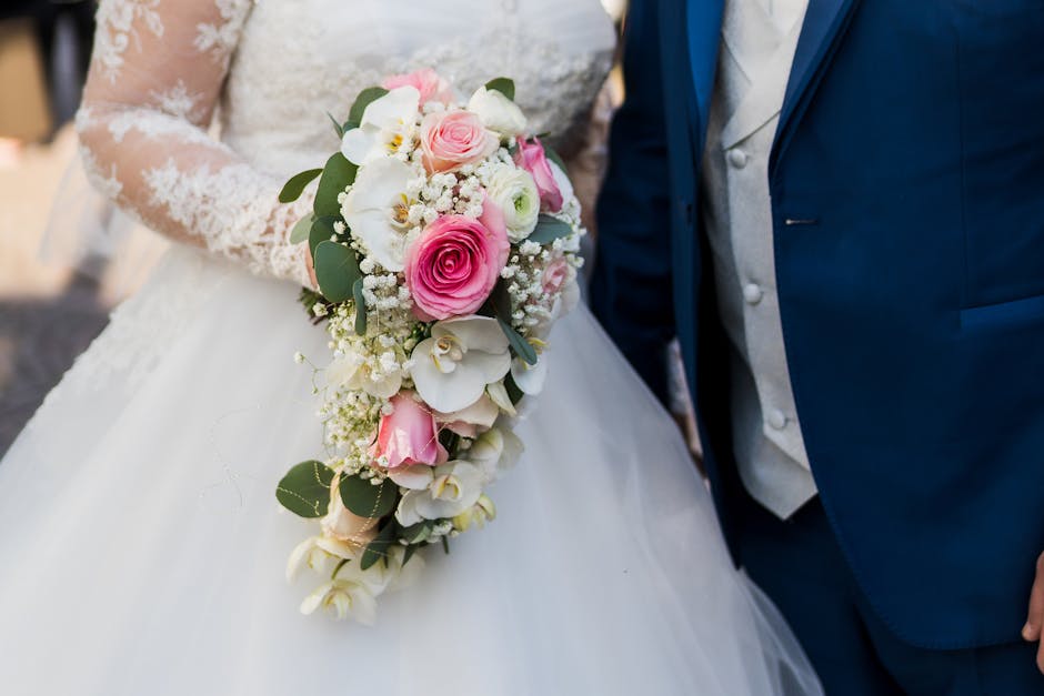 A wedding bouquet arrangement featuring pink roses, white orchids, and small white filler flowers, accented with green eucalyptus leaves. The bouquet is held by a bride dressed in a white lace wedding