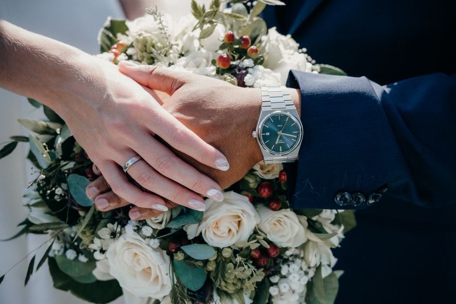 Close-up of a bride and groom's hands gently clasped over a bouquet of fresh flowers, including white roses, green foliage, and small red berries. The bride's hand features a wedding band and neatly m