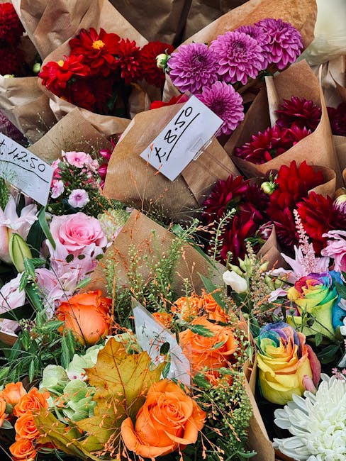 A vibrant assortment of freshly cut flowers arranged in individual paper wraps, prominently featuring red dahlias, pink chrysanthemums, purple asters, orange roses, rainbow-colored roses, and white ch