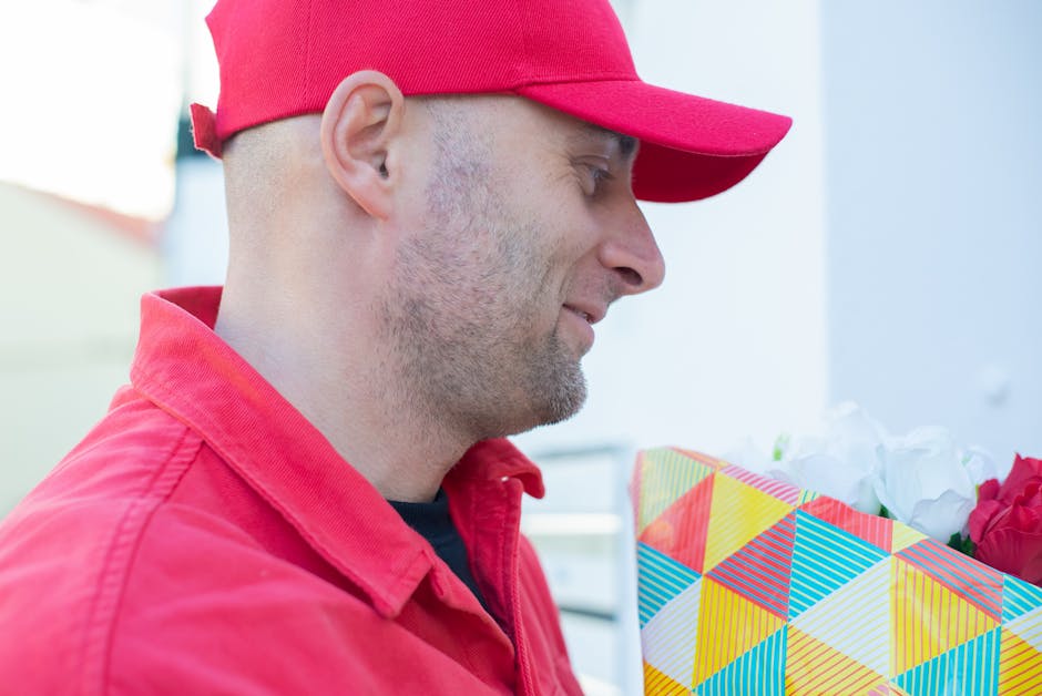A man dressed in a red cap and red jacket is seen in profile, holding a wrapped floral gift with vibrant, geometric-patterned wrapping paper featuring red, yellow, blue, and white triangles. The bouqu